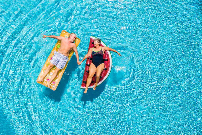 Above vertical view of people old senior couple taking hands with love and having fun on the blue clear swimming pool together enjoying the summer holiday vacation with trendy coloured lilos inflatable mattress