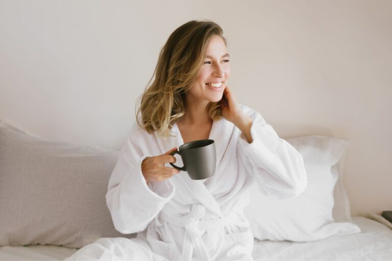 Young beautiful woman wearing white bathrobe having breakfast in bed with coffee and croissant and fresh fruits in cozy bedroom. Morning rituals.