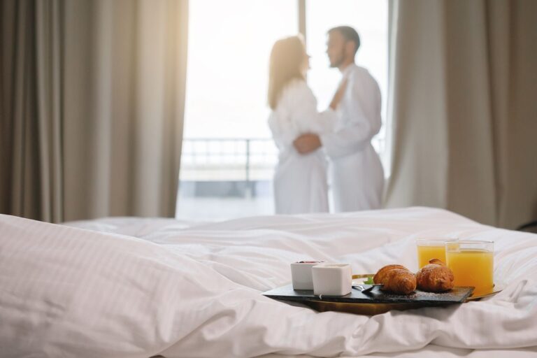 Breakfast tray is on a white bed. A couple in bathrobes are drinking coffee by the window in the background. Selective focus.